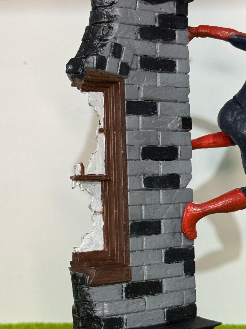 Op de afbeelding: Gedetailleerd beeldje van een bakstenen muur met een gebroken raam. De muur is grijs en zwart geschilderd, met een bruin kozijn. Een figuur in een rood en blauw pak beklimt de muur.