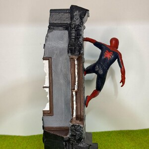 Op de afbeelding: Gedetailleerd beeldje van Spider-Man in een dynamische pose, vastklampend aan een verweerde gevel. Het figuur draagt een klassiek rood-zwart pak, het gebouw heeft grijs en zwart metselwerk en een raam.