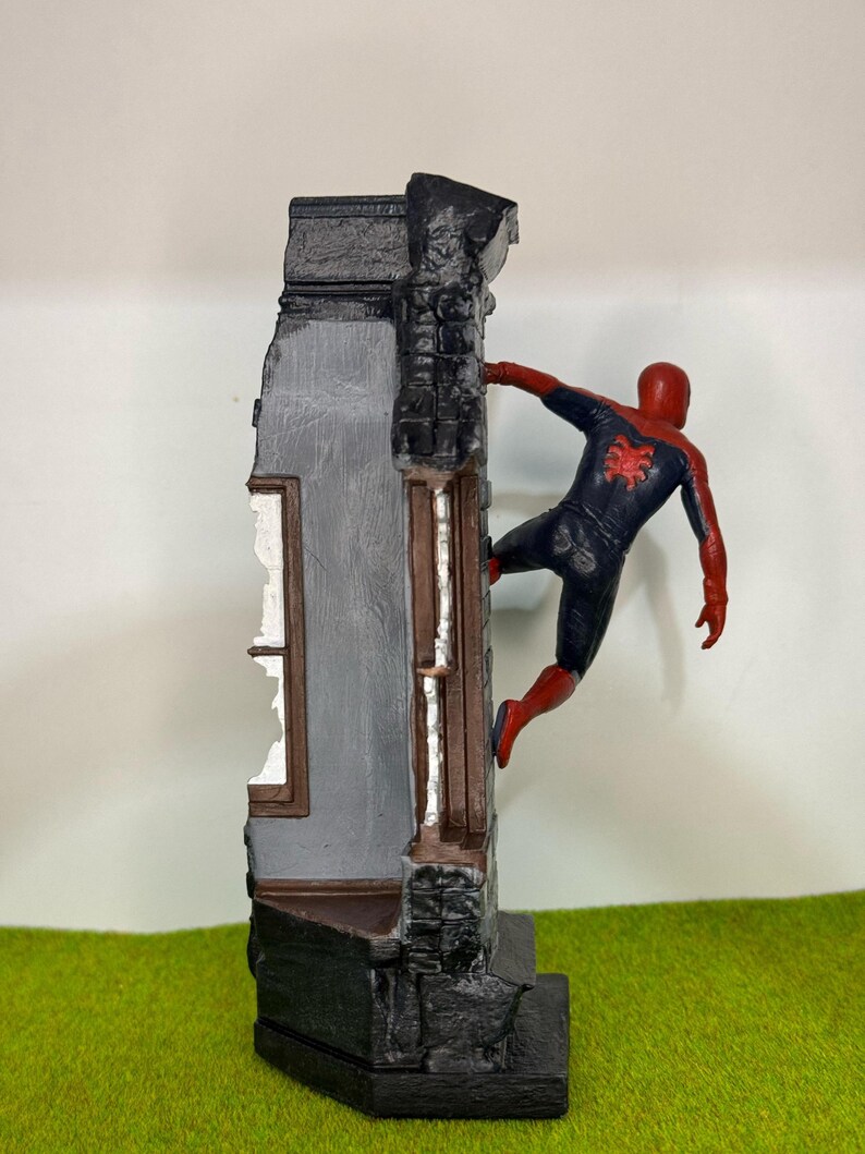 Op de afbeelding: Gedetailleerd beeldje van Spider-Man in een dynamische pose, vastklampend aan een verweerde gevel. Het figuur draagt een klassiek rood-zwart pak, het gebouw heeft grijs en zwart metselwerk en een raam.
