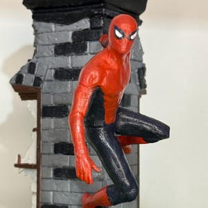Op de afbeelding: Een rood en zwart Spider-Man beeldje dat zich vastklampt aan een grijze bakstenen muur. De figuur staat in een dynamische pose, met &eacute;&eacute;n been uitgestrekt en armen uitgestrekt. De muur heeft een zwarte bovenkant en een gedeeltelijk verwoest gedeelte.
