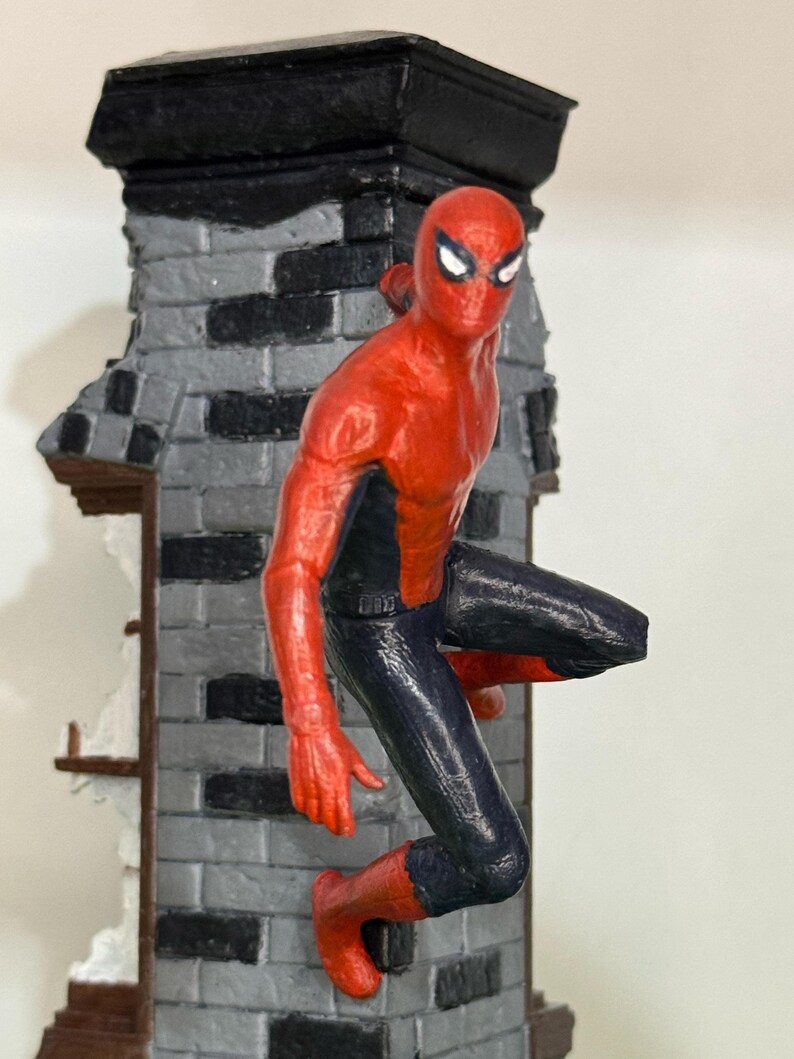 Op de afbeelding: Een rood en zwart Spider-Man beeldje dat zich vastklampt aan een grijze bakstenen muur. De figuur staat in een dynamische pose, met &eacute;&eacute;n been uitgestrekt en armen uitgestrekt. De muur heeft een zwarte bovenkant en een gedeeltelijk verwoest gedeelte.