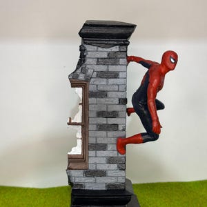 Op de afbeelding: Een beeldje van Spider-Man in een rood en zwart pak, dat zich vastklampt aan een grijze bakstenen muur met een gebroken raam. De muur heeft een zwarte basis en bovenkant, tegen een groene achtergrond.