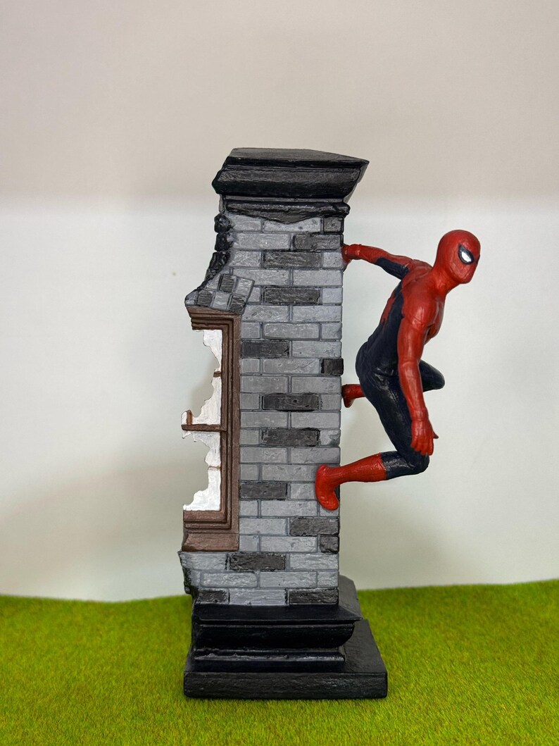 Op de afbeelding: Een beeldje van Spider-Man in een rood en zwart pak, dat zich vastklampt aan een grijze bakstenen muur met een gebroken raam. De muur heeft een zwarte basis en bovenkant, tegen een groene achtergrond.