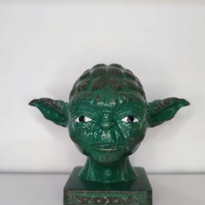 May include: Green Yoda figurine with a distressed paint finish. The figurine is on a square base with the word "Yoda" inscribed on the front.