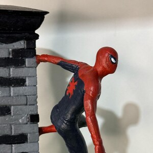 Op de afbeelding: Een rood en marineblauw Spider-Man beeldje dat een grijze bakstenen muur beklimt. Het beeldje heeft een rood spinnenembleem op de borst en witte ogen. De muur heeft een zwarte bovenkant.