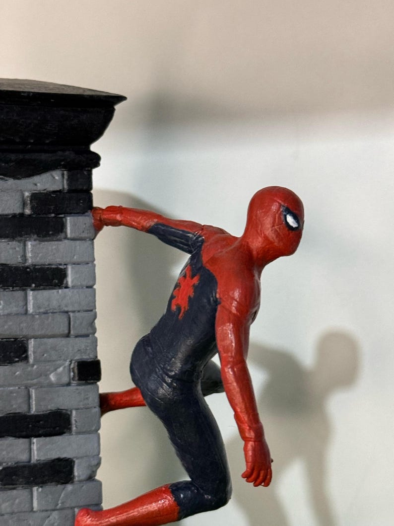 Op de afbeelding: Een rood en marineblauw Spider-Man beeldje dat een grijze bakstenen muur beklimt. Het beeldje heeft een rood spinnenembleem op de borst en witte ogen. De muur heeft een zwarte bovenkant.