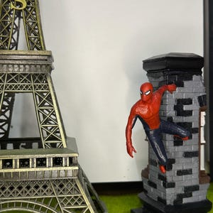 Op de afbeelding: Een miniatuur Eiffeltoren en een Spider-Man beeldje dat zich vastklampt aan een bakstenen structuur. De Eiffeltoren is bronskleurig en het Spider-Man figuur is rood en blauw. De bakstenen structuur is grijs en zwart.