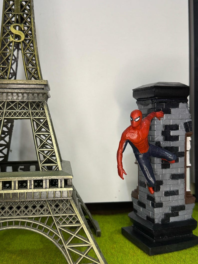 Op de afbeelding: Een miniatuur Eiffeltoren en een Spider-Man beeldje dat zich vastklampt aan een bakstenen structuur. De Eiffeltoren is bronskleurig en het Spider-Man figuur is rood en blauw. De bakstenen structuur is grijs en zwart.