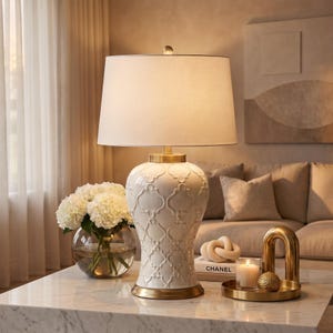 May include: A cream-colored ceramic table lamp with a white lampshade. The lamp has a gold base and a decorative pattern. A vase of white flowers and decorative items sit on a marble table.