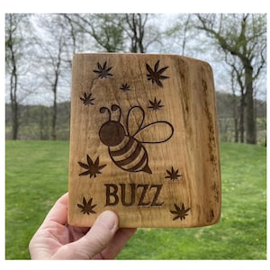 May include: A wooden block with a carved bee design and the word "BUZZ". The bee is surrounded by carved leaf shapes. The block is held in a hand against a background of green grass and trees.