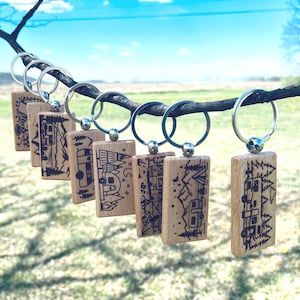 May include: A set of eight wooden keychains with a camping theme. Each keychain features a different design of a camper van, trees, and mountains. The keychains are attached to silver key rings.