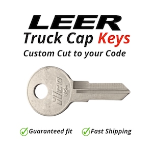 May include: A silver-colored truck cap key with the text "LEER Truck Cap Keys Custom Cut to your Code". The key has a circular head with a hole and the text "Ilco USA".