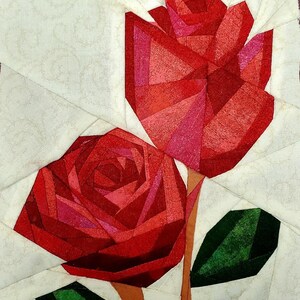 Könnte beinhalten: Ein Stoff-Quiltblock mit zwei stilisierten Rosen, die aus geometrischen Stücken aus rotem und rosafarbenem Stoff zusammengesetzt sind. Die Rosen haben lange braune Stiele und dunkelgrüne Blätter auf einem cremefarbenen Hintergrund mit Wirbelmuster.