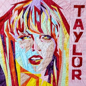 Puede incluir: Una colcha de retazos colorida con un retrato de una mujer con cabello rojo y una camisa con estampado rojo, blanco y azul. La colcha está hecha de varias telas y texturas, creando un diseño único y artístico. La palabra "TAYLOR" está cosida en rojo en el lado derecho de la colcha.