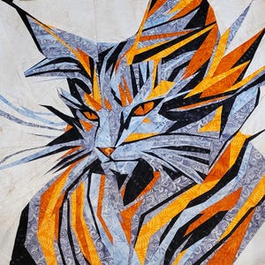 以下が含まれることがあります： 猫の顔を表現したカラフルなパッチワークキルトのクローズアップ。キルトは、青、オレンジ、黒のさまざまな色合いの生地で作られており、幾何学的なパターンを生み出しています。猫の目は鮮やかな緑色です。