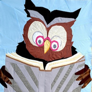 Könnte beinhalten: Ein Stoff-Quiltblock mit einer braunen Eule mit großen weißen Augen, rosa Pupillen und einem gelben Schnabel, die ein graues Buch mit sichtbaren Seiten liest. Die Eule ist vor einem hellblauen Hintergrund mit geometrischen Stoffteilen.