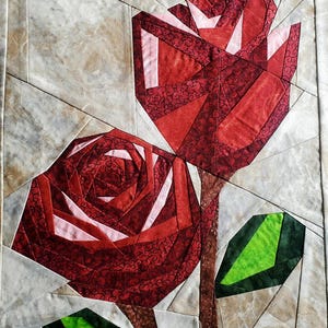 Könnte beinhalten: Ein Stoff-Quiltblock mit zwei Rosen, die aus geometrischen Stücken aus rotem, dunkelrotem und hellrosa gemustertem Stoff zusammengesetzt sind. Die Rosen haben braune Stiele und hellgrüne Blätter auf einem Hintergrund aus beigem Stoff.