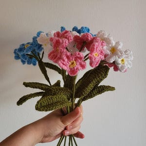 Peut inclure: Un bouquet de fleurs crochetées en rose, blanc et bleu. Les fleurs sont disposées en bouquet tenu à la main avec des tiges et des feuilles crochetées vertes.