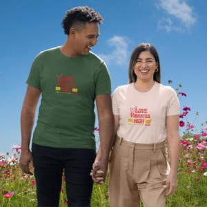 May include: Two people wearing matching t-shirts with the text "Love Vibrates High" in red and yellow. The shirts are olive green and cream-colored. The couple is holding hands in front of a blue sky and a field of flowers.