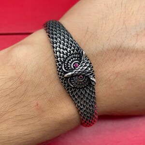 May include: Silver-toned owl bracelet with intricate feather detailing. The owl's eyes are accented with small, round, red gemstones. The bracelet is set against a red background, showcasing the jewelry's design.