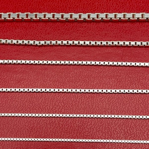 May include: Several silver box chain necklaces of varying thicknesses are displayed against a red background. The chains are made of small, rectangular links, creating a sleek and modern look. The chains are perfect for layering or wearing alone.