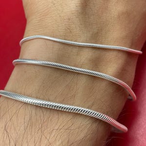 Puede incluir: Tres pulseras de cadena de serpiente plateadas se usan en una muñeca. Las pulseras están hechas de un material metálico brillante y tienen un patrón texturizado en forma de serpiente. Las pulseras están apiladas una encima de la otra, creando un aspecto en capas.