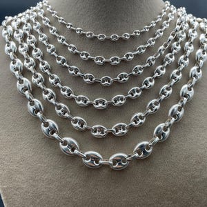 Op de afbeelding: Een display van zes zilverkleurige kettingkettingen met een boonvormig schakelontwerp. De kettingen variëren in lengte en tonen de verschillende beschikbare maten. De sieraden worden gepresenteerd op een mannequin buste in neutrale tinten.