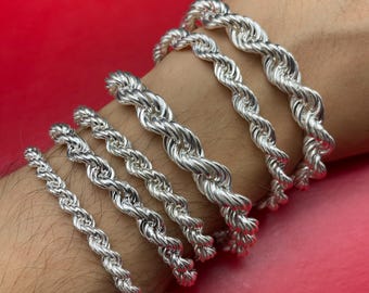 Pode incluir: Uma coleção de pulseiras de corrente de corda prateadas em várias espessuras. As pulseiras são exibidas no pulso e braço de uma pessoa contra um fundo vermelho. As pulseiras têm um design torcido.