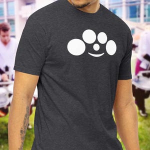 Può includere: Una t-shirt grigio scuro melange con una grafica bianca di cinque piatti di batteria disposti a semicerchio. I piatti sono disposti in modo da assomigliare a un viso sorridente.