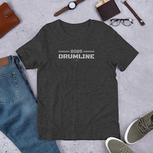 Può includere: T-shirt grigio scuro con la scritta "2025 DRUMLINE" in lettere bianche. La maglietta è a maniche corte e ha lo scollo tondo. La maglietta è appoggiata su una superficie grigio chiaro con altri accessori.