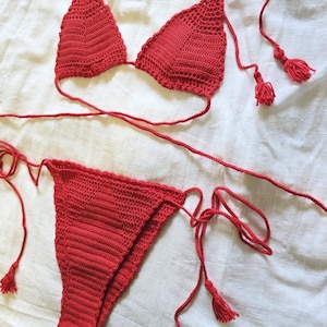 Può includere: Un bikini rosso all'uncinetto fatto a mano. Il top è a triangolo con lacci regolabili. Lo slip è un classico modello bikini con lacci abbinati. Il set include nappe sui lacci. Il bikini è esposto su una superficie bianca.