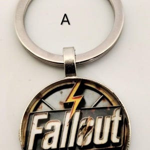 May include: A round keychain with a silver ring and a Fallout-themed pendant. The pendant features the word "Fallout" in white letters, a yellow lightning bolt, and a biohazard symbol. The design has a distressed, rusty appearance.