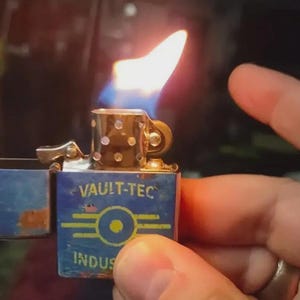 May include: A lit, vintage-style lighter with a blue and yellow Vault-Tec logo and text. The lighter has a flame with blue and yellow hues. The lighter is open, revealing the flint and wick.