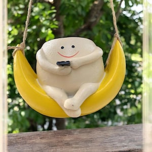 Op de afbeelding: Een wit keramiek beeldje van een lachend persoon die in een gele halve maan plantenpot zit. Het beeldje heeft armen en benen en houdt een klein zwart voorwerp in zijn handen.