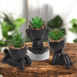 May include: Three black ceramic planters shaped like sitting figures, each with a different succulent plant inside. The planters are all facing forward and are arranged on a wooden surface.