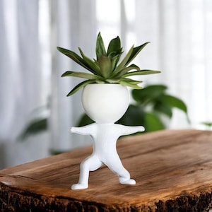 Puede incluir: Una maceta de cerámica blanca con forma de persona en postura de yoga, sosteniendo una planta suculenta verde. La maceta tiene una cabeza redonda y brazos extendidos. La planta tiene hojas verdes con puntas rojizas.