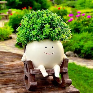 May include: A round, off-white planter shaped like a character with a simple smiling face, seated in a small brown rocking chair. The planter is filled with a dense, vibrant green artificial plant with small, rounded leaves and a bushy texture.