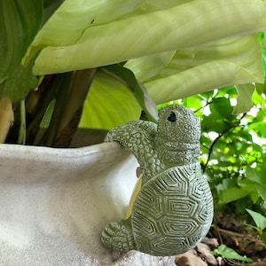 May include: A green ceramic turtle figurine with a textured shell, climbing up the side of a white ceramic planter. The turtle's head is turned to the side, and its eyes are black.