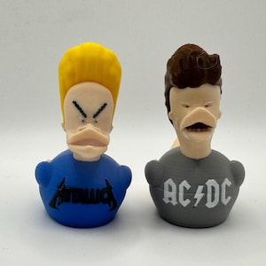 May include: Two novelty rubber ducks, each with a cartoon character design. One duck has yellow hair and a blue shirt with "METALLICA" printed on it. The other duck has brown hair and a gray shirt with "AC/DC" printed on it.