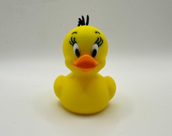 May include: A yellow rubber duck toy with a cartoonish design. The duck has a bright orange beak, black eyes, and black hair. The duck is a classic bath toy.