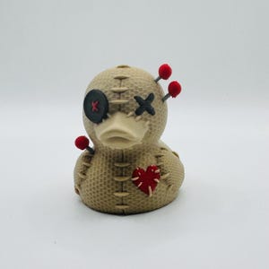 May include: A beige voodoo doll duck with stitched details, a black button eye, and a stitched cross eye. It has a red heart patch and red pin accents, creating a unique and quirky decorative item.