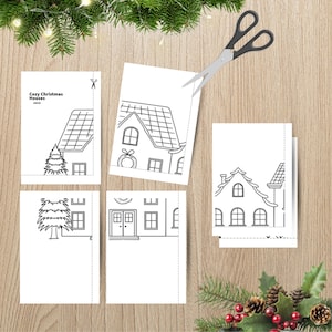May include: A set of white paper sheets featuring black line art of houses and Christmas trees with the text Cozy Christmas Houses 100CM. The sheets include dashed cutting lines and are accompanied by a pair of black metal scissors.