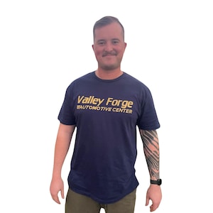 May include: A navy blue t-shirt with the text "Valley Forge Automotive Center" in gold lettering. The person is wearing a black watch and has a tattoo on their arm. The background is white.