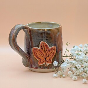 May include: A handmade ceramic mug with a leaf design. The mug features a brown, blue, and orange color scheme. The handle is a curved shape. The mug is decorated with an orange leaf. White flowers are in the background.