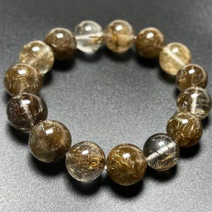 May include: A bracelet made of clear, round beads with golden-brown inclusions. The beads are strung together, forming a circular shape. The bracelet is displayed on a dark surface, highlighting the natural colors and textures of the stones.
