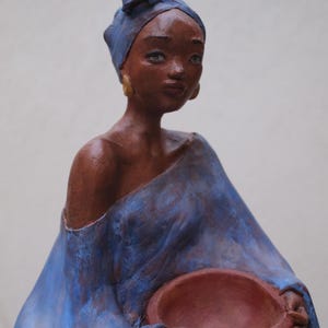 Peut inclure: Une figurine sculptée représentant une personne à la peau brun foncé, portant un foulard bleu et un vêtement bleu dénudant une épaule. La figurine porte des boucles d'oreilles créoles dorées et tient un bol rond brun-rouge simple.