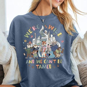 Puede incluir: Una camiseta azul con el texto "WE'RE WILD AND WE CAN'T BE TAMED" y un gr&aacute;fico colorido de personajes animales frente a un horizonte urbano. La camiseta es llevada.