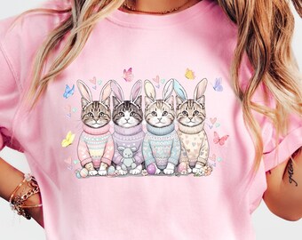 May include: A light pink t-shirt featuring a watercolor illustration of four cats wearing bunny ears and sweaters. The cats are surrounded by butterflies. The shirt is a crew neck and has short sleeves.
