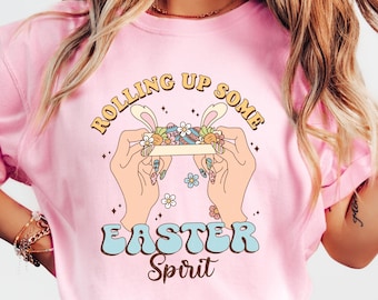 May include: A pink t-shirt with the text "Rolling Up Some Easter Spirit" in a retro font. The design features two hands rolling a joint with Easter eggs and bunny ears.