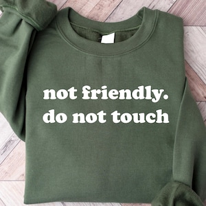 May include: An olive green sweatshirt with the text "not friendly. do not touch" in white. The sweatshirt has long sleeves and a crew neck. The garment is displayed on a light wood grain surface.